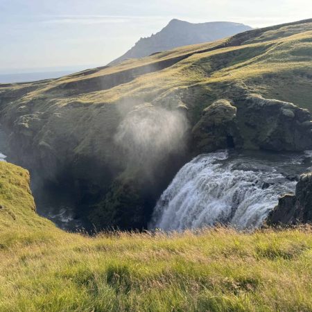 Scenic Icelandic waterfall with lush green hills and mountain backdrop.